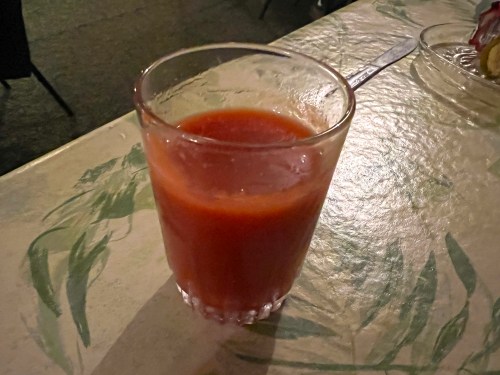 A glass of red beverage sits on a table with a floral design, illuminated by soft lighting.