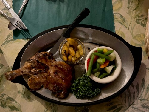 A plate with grilled chicken, sautéed vegetables, and a small bowl of fruit, set on a floral tablecloth.