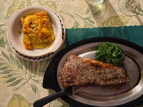 A plated meal featuring a large steak with a side of cheesy potato casserole and a garnish of kale.