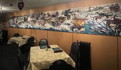 A cozy restaurant interior featuring tables and a large mural depicting a winter scene with sled dogs and people.