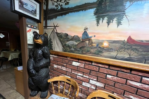 A wooden bear statue stands beside a mural of a camper by a fire, surrounded by trees and a canoe. Cozy interior setting.