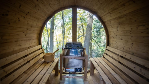 Interior of a wooden sauna with benches, a stove, and large circular windows overlooking a forest.