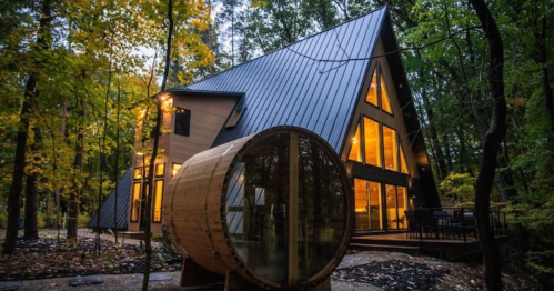 A modern A-frame cabin surrounded by trees, featuring large windows and a unique round sauna in the foreground.