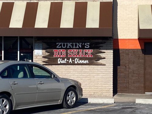 Sign for Zukin's Rib Shack, featuring "Dial-A-Dinner," with a parked car in front of the restaurant.
