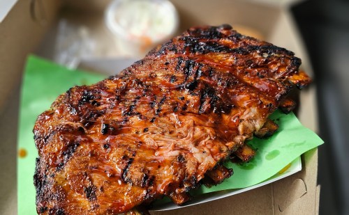A deliciously grilled rack of ribs on a green paper, accompanied by a small container of coleslaw.