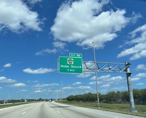 Green highway sign indicating "Exit 96 Hobe Sound 1 Mile" with a clear blue sky and clouds in the background.