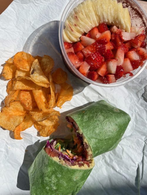 A green wrap filled with vegetables, a side of orange chips, and a bowl of fruit with strawberries and pineapple.