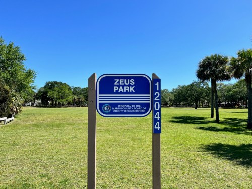Sign for Zeus Park, featuring blue and white colors, with a clear blue sky and green grass in the background.
