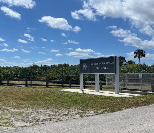 Sign for Jonathan Dickinson State Park with a blue sky and scattered clouds in the background. Green vegetation nearby.