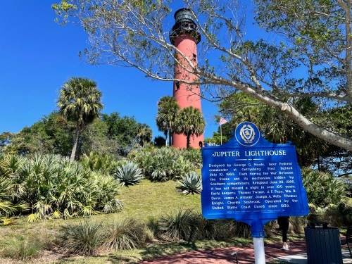 A red and black lighthouse surrounded by palm trees, with a historical sign in front detailing its history.