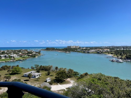 A scenic view of a blue river winding through lush greenery, with buildings and the ocean in the background under a clear sky.