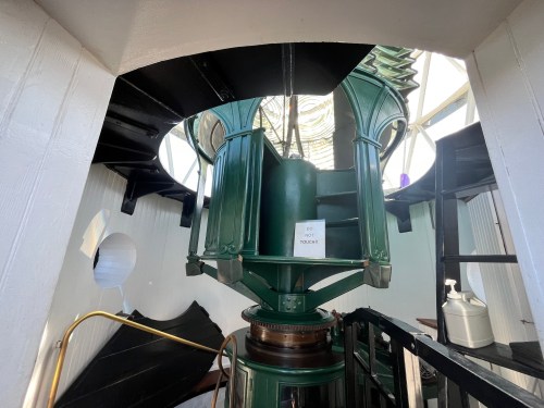 Interior view of a lighthouse lantern room with a green light and a sign that says "Do Not Touch." Spiral staircase visible.
