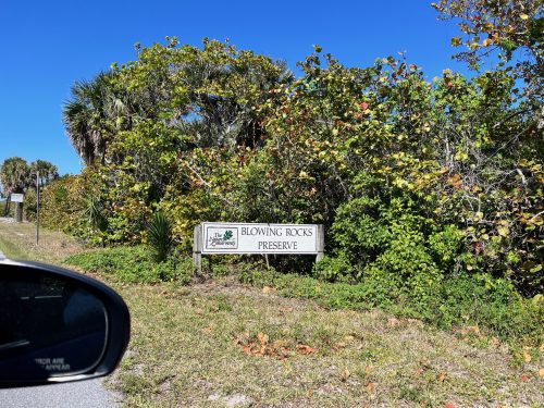 Sign for Blowing Rocks Preserve surrounded by lush greenery and blue sky, viewed from a car.