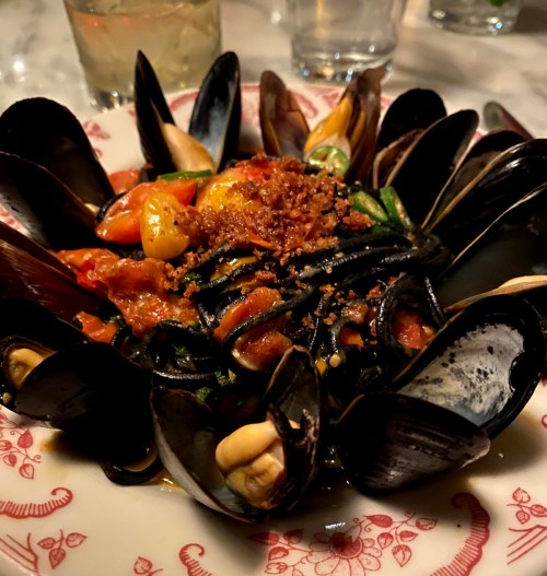 A plate of black pasta topped with mussels, tomatoes, and crispy bits, garnished with herbs.