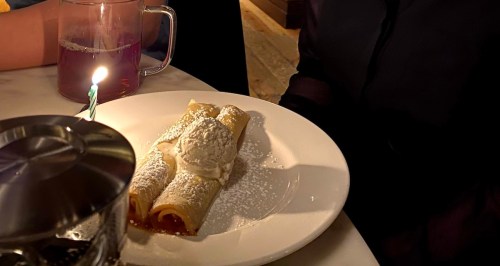 A plate with two crepes topped with whipped cream and powdered sugar, accompanied by a drink and a birthday candle.