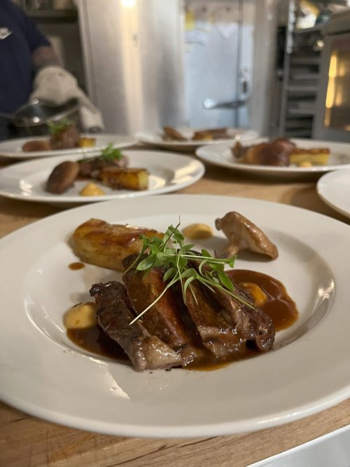 A plated dish featuring sliced meat, sauce, roasted potatoes, and garnished with microgreens, set in a kitchen.