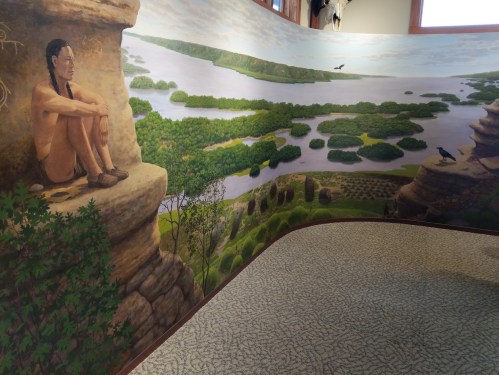 A mural depicting a serene landscape with a figure sitting on a rock, surrounded by lush greenery and a river.