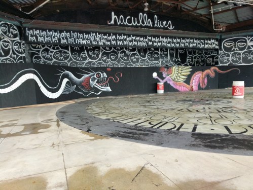 A mural featuring a snake and a colorful dragon, with stylized faces and text on a black wall in an open space.