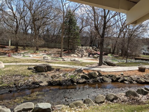 A serene park scene featuring a stream, rocky paths, and trees, with a small hill in the background.
