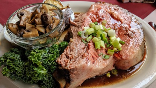 A plate featuring a large slice of roast beef with green onions, served with mushrooms and a baked potato on the side.