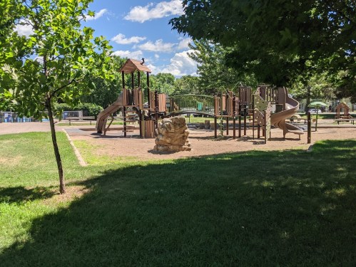 A playground with slides and climbing structures surrounded by trees and grassy areas under a blue sky.