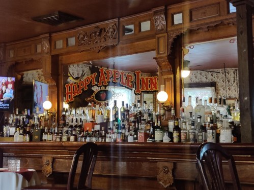 A wooden bar with a variety of liquor bottles displayed, featuring a sign that reads "Happy Apple Inn" in the background.