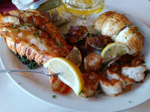 A plate of grilled seafood featuring lobster, fish, scallops, and garnished with lemon and herbs.