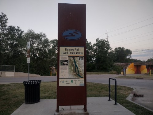 Sign for Phinney Park Lizard Creek Access, featuring park information and a map, surrounded by greenery.