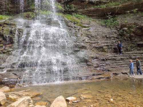 A waterfall cascades down rocky cliffs, with two people walking along the water's edge and one person standing nearby.