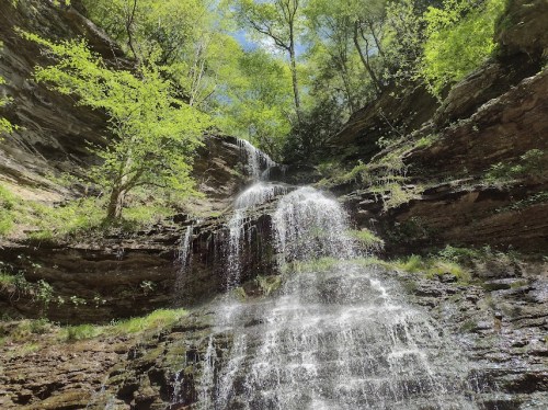 A serene waterfall cascading down rocky cliffs, surrounded by lush green trees and bright blue sky.