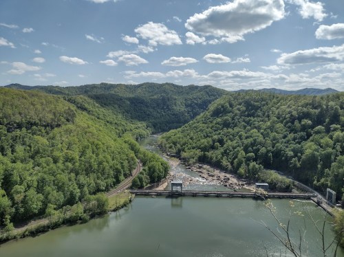 A scenic view of a river and dam surrounded by lush green hills under a partly cloudy sky.