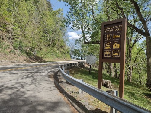 A scenic view of a road leading to Hawks Nest State Park, featuring a sign with park information and nearby greenery.