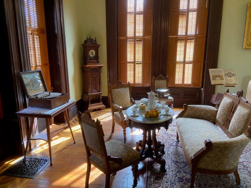 A vintage sitting room with ornate furniture, a clock, and a tea set on a table, illuminated by sunlight through windows.
