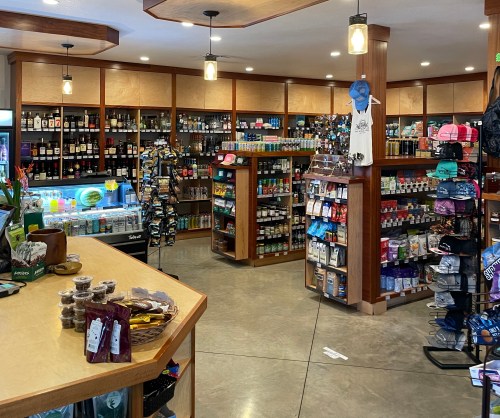 A well-stocked store interior featuring shelves of snacks, drinks, and merchandise, with a wooden counter in the foreground.