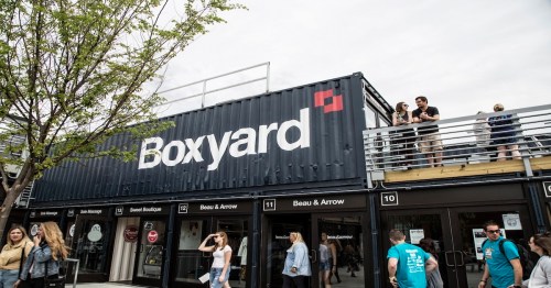 A shipping container building with "Boxyard" signage, people on the rooftop, and visitors exploring shops below.