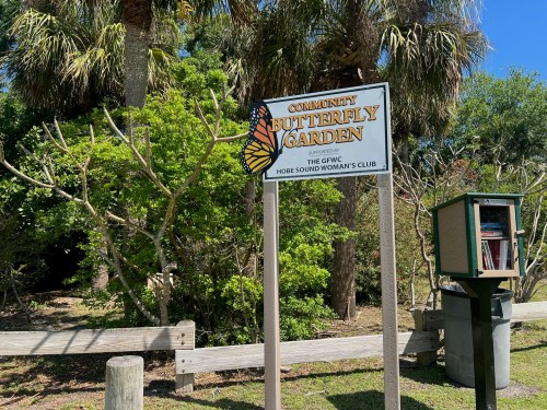 Sign for the Community Butterfly Garden, surrounded by greenery and palm trees, with a small library box nearby.
