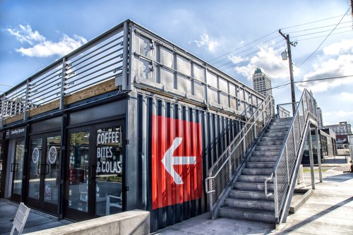 A modern building made from shipping containers, featuring a large red arrow and a staircase leading to the upper level.