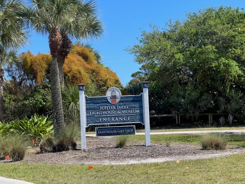 Sign for Jupiter Inlet Lighthouse & Museum entrance, featuring palm trees and a clear blue sky.