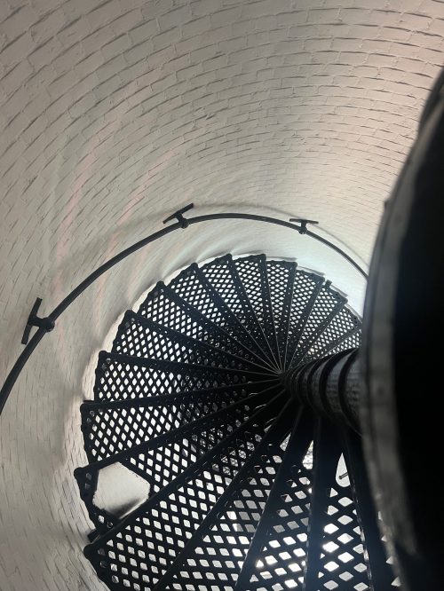 A spiral staircase viewed from above, featuring a black metal railing and textured steps against a white brick wall.