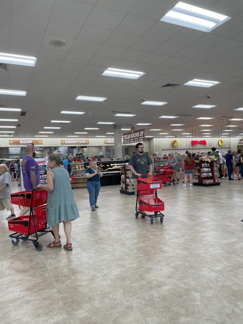 A busy store interior with shoppers holding red baskets, browsing various food items and displays.