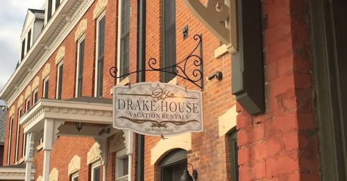 Sign for "Drake House Vacation Rentals" hanging from a building with red brick and decorative architecture.