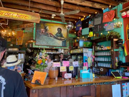 A vibrant café interior with colorful decor, a large mural, and a busy counter filled with drinks and merchandise.