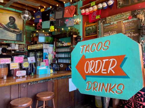Colorful bar interior with a sign reading "TACOS ORDER DRINKS" and various hats and decorations hanging above.