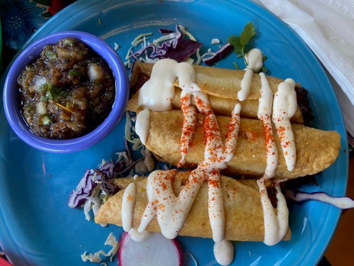 A blue plate with three crispy tacos drizzled with sauce, accompanied by a small bowl of salsa and shredded cabbage.