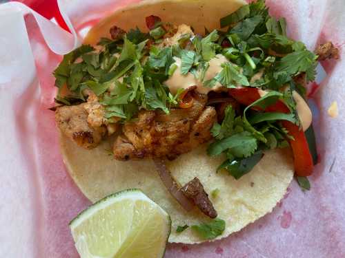 A taco filled with grilled meat, topped with cilantro, peppers, and sauce, served with a lime wedge on the side.