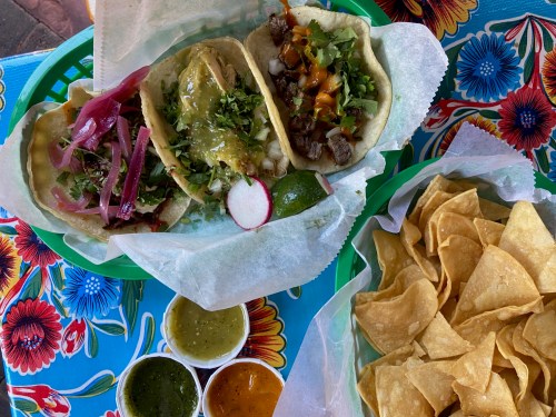 Three tacos with various toppings and a side of tortilla chips and three dipping sauces on a colorful floral tablecloth.