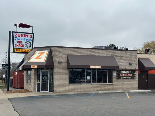 Exterior of Zukin's Rib Shack and Pizza, featuring a sign and a modern building with a brown awning.