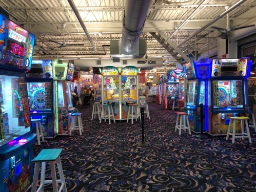 A vibrant arcade filled with various game machines and colorful lights, featuring a patterned carpet and seating areas.
