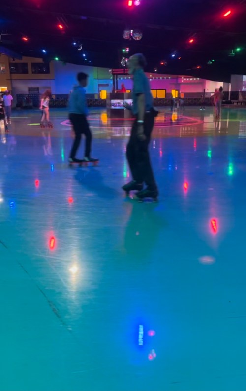 Two people skate on a brightly lit roller rink with colorful lights reflecting on the smooth floor.
