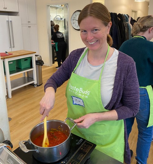 A smiling woman in an apron stirs a pot of sauce in a cooking class, with others working in the background.